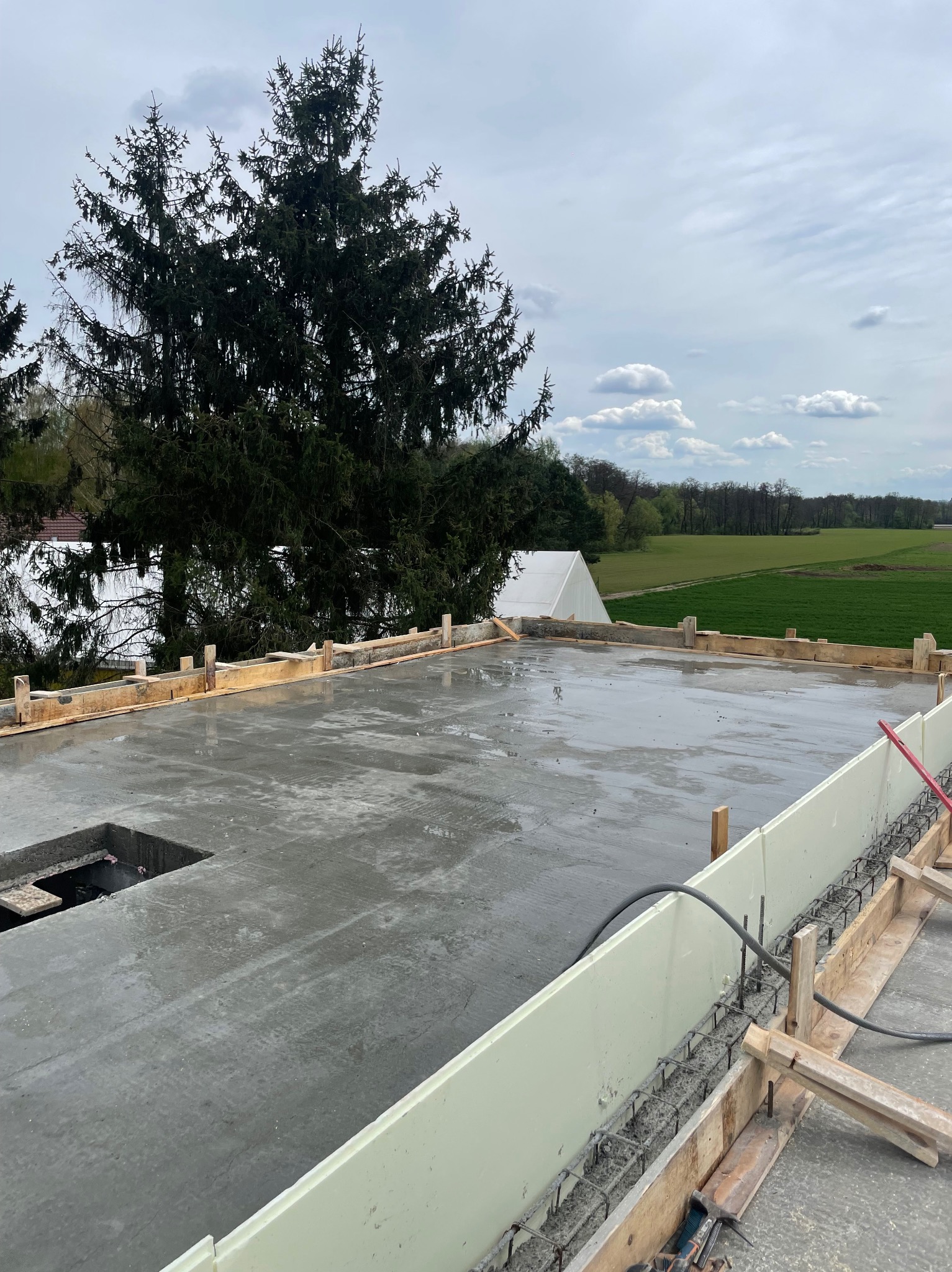 Zalany betonowy fundament z widocznym zbrojeniem i szalunkiem, na tle zieleni i pochmurnego nieba. Widoczny otwór w fundamencie.