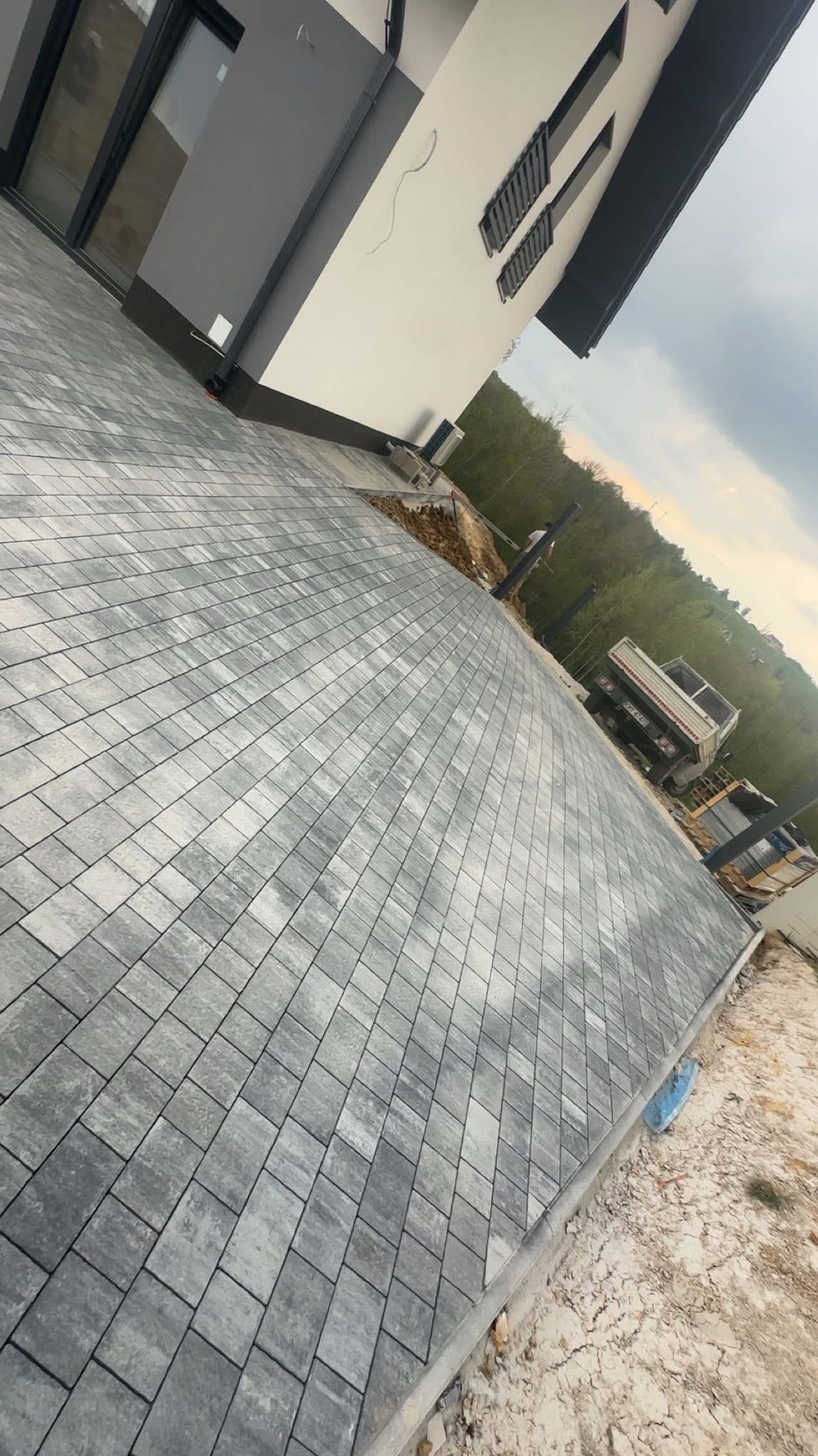 Ułożona szara kostka brukowa na podjeździe domu z szarą elewacją. W tle widoczny samochód ciężarowy i drzewa. Ujęcie z niskiej perspektywy.