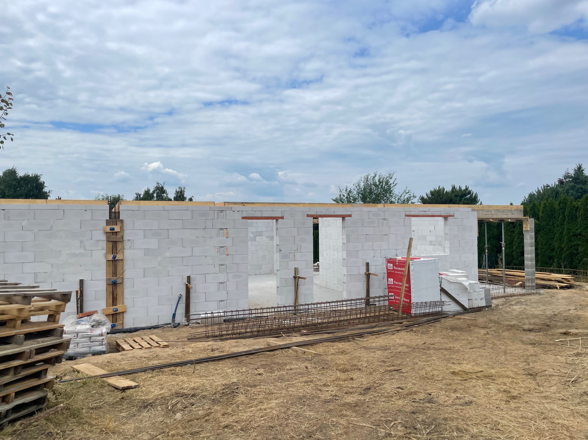 Wznoszenie ścian domu z betonu komórkowego. Widoczne zbrojenia, palety z materiałami budowlanymi i elementy drewniane. Etap budowy w stanie surowym otwartym.