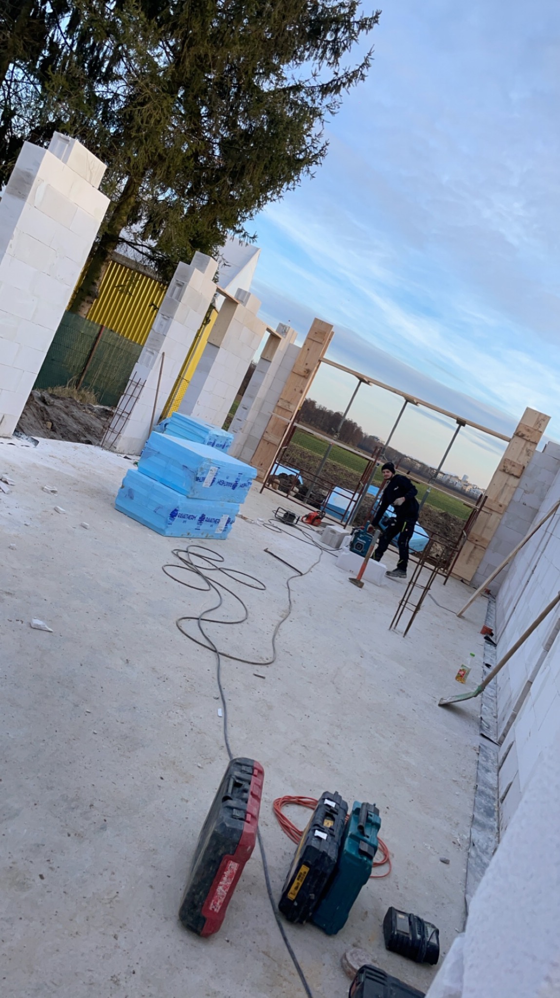 Budowa domu z białych bloczków: pracownik z narzędziem, widoczne materiały izolacyjne i narzędzia. Etap wznoszenia ścian, częściowo zbrojonych.