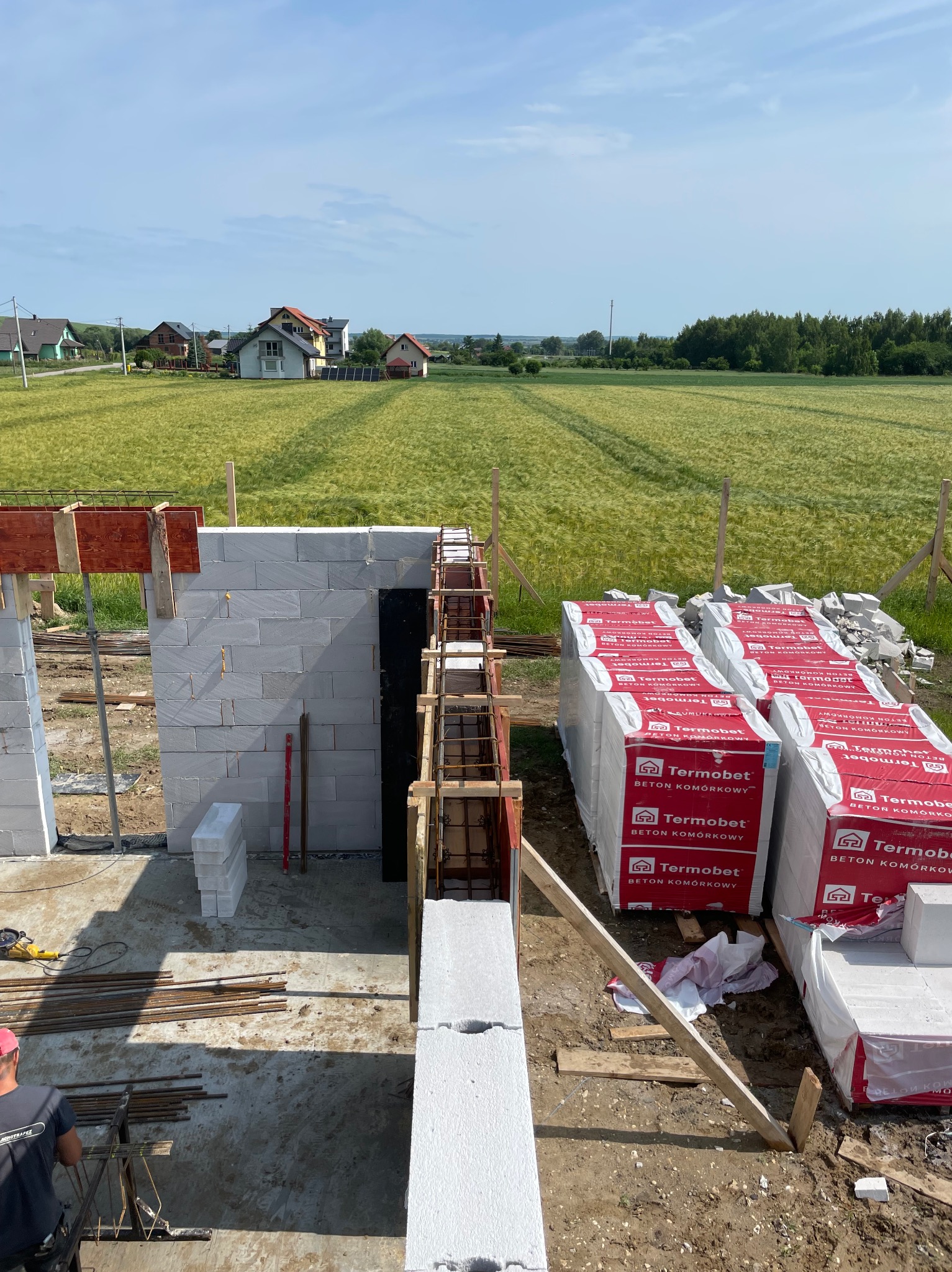 Wznoszenie ścian domu z betonu komórkowego Termobet. Widoczne zbrojenia, szalunki i narzędzia. W tle zielone pola i zabudowa wiejska.