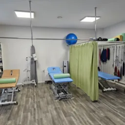 Jasne wnętrze gabinetu rehabilitacyjnego z trzema profesjonalnymi stanowiskami do terapii manualnej, każde wyposażone w regulowany stół, wałek oraz dodatkowy sprzęt medyczny, widoczne zasłony...