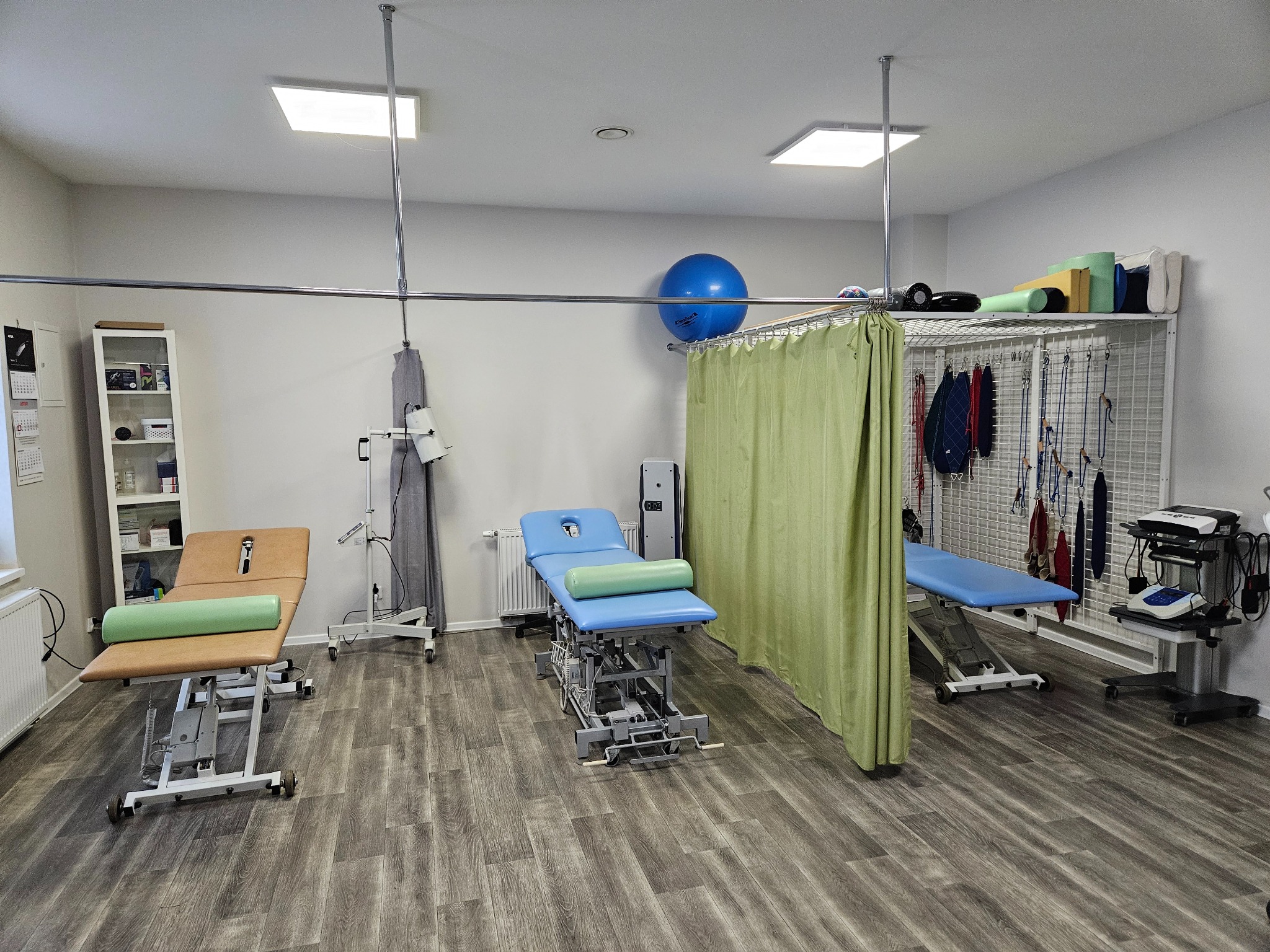 Jasne wnętrze gabinetu rehabilitacyjnego z trzema profesjonalnymi stanowiskami do terapii manualnej, każde wyposażone w regulowany stół, wałek oraz dodatkowy sprzęt medyczny, widoczne zasłony...