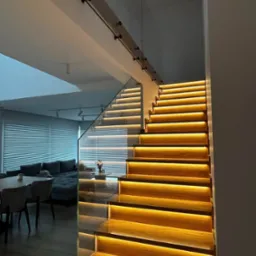 Nowoczesne schody z podświetlanymi stopniami i szklaną balustradą, widok z dołu na tle salonu z szarą sofą i żaluzjami.