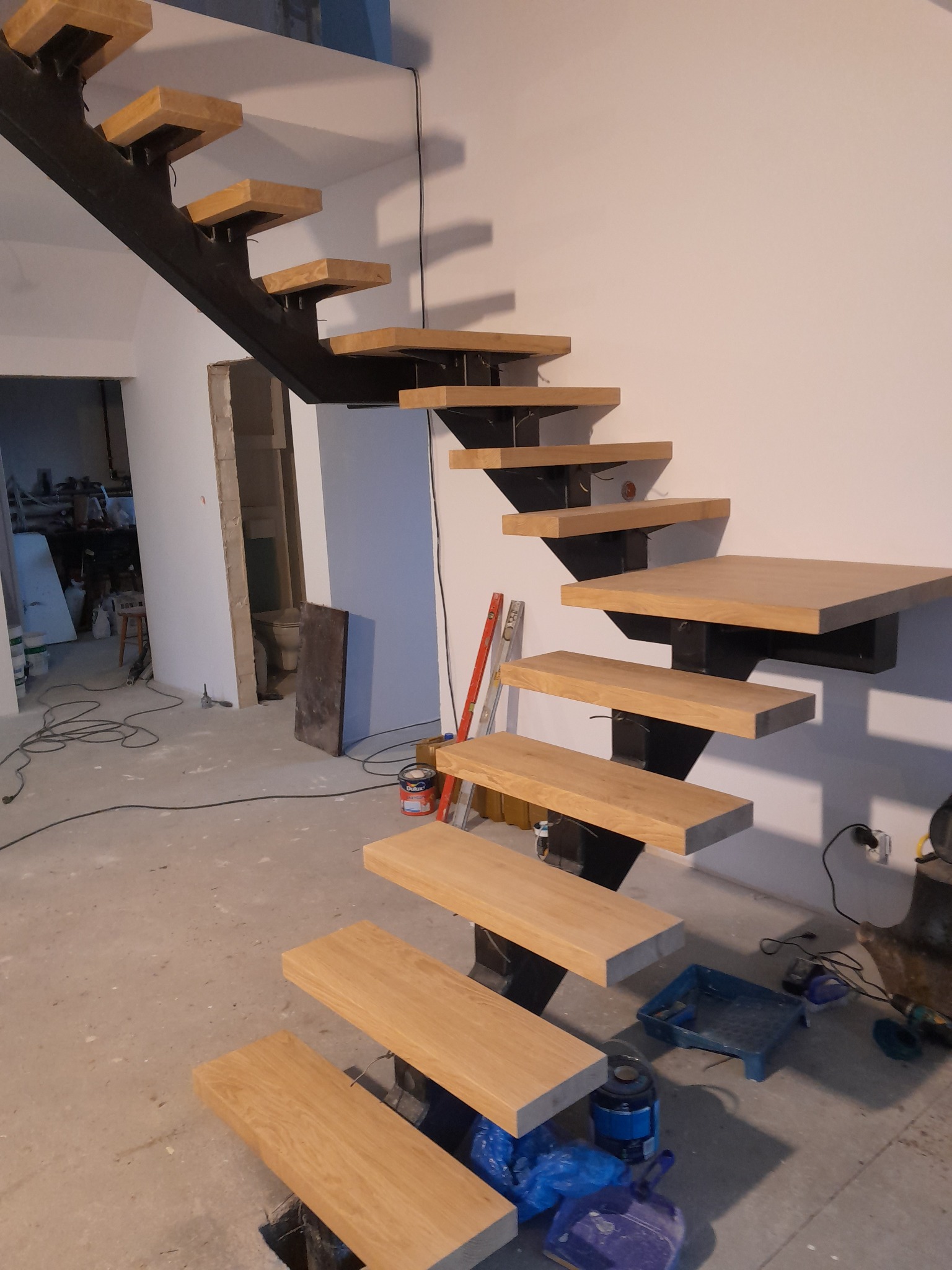 Ażurowe schody z drewnianymi stopniami i czarną metalową konstrukcją w trakcie montażu, widoczne narzędzia i materiały budowlane w surowym wnętrzu.