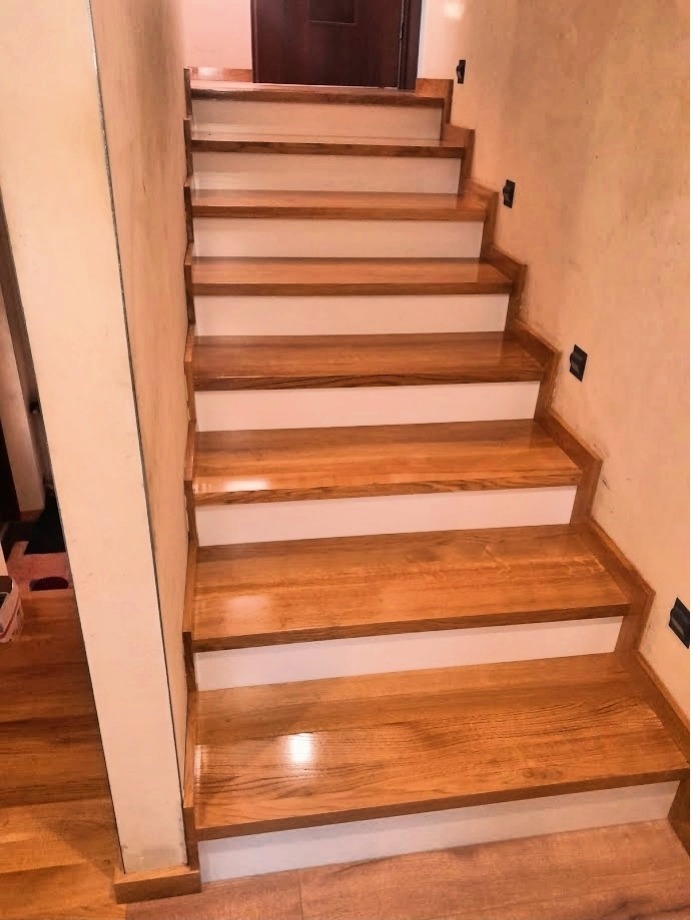 Wykończone schody z drewnianymi stopniami o ciepłym odcieniu i białymi podstopnicami, widoczne czarne punkty instalacji oświetleniowej na ścianie.