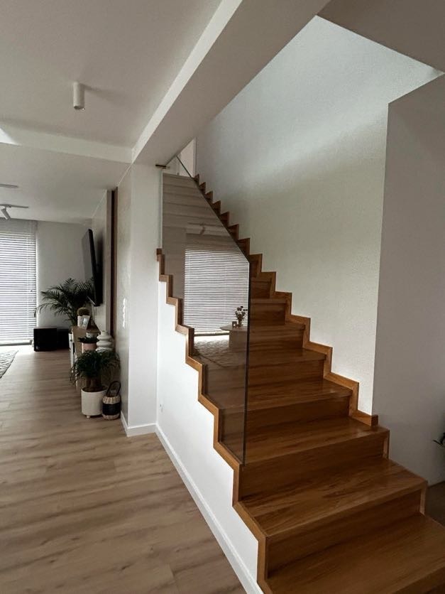 Nowoczesne schody z drewna o ciepłym odcieniu, z przezroczystą szklaną balustradą, prowadzące do jasnego pomieszczenia z roletami i roślinami doniczkowymi.