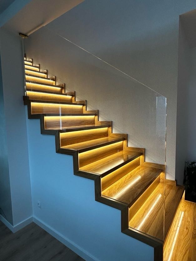 Nowoczesne schody drewniane z podświetleniem LED pod każdym stopniem i szklaną balustradą, widok z dołu.