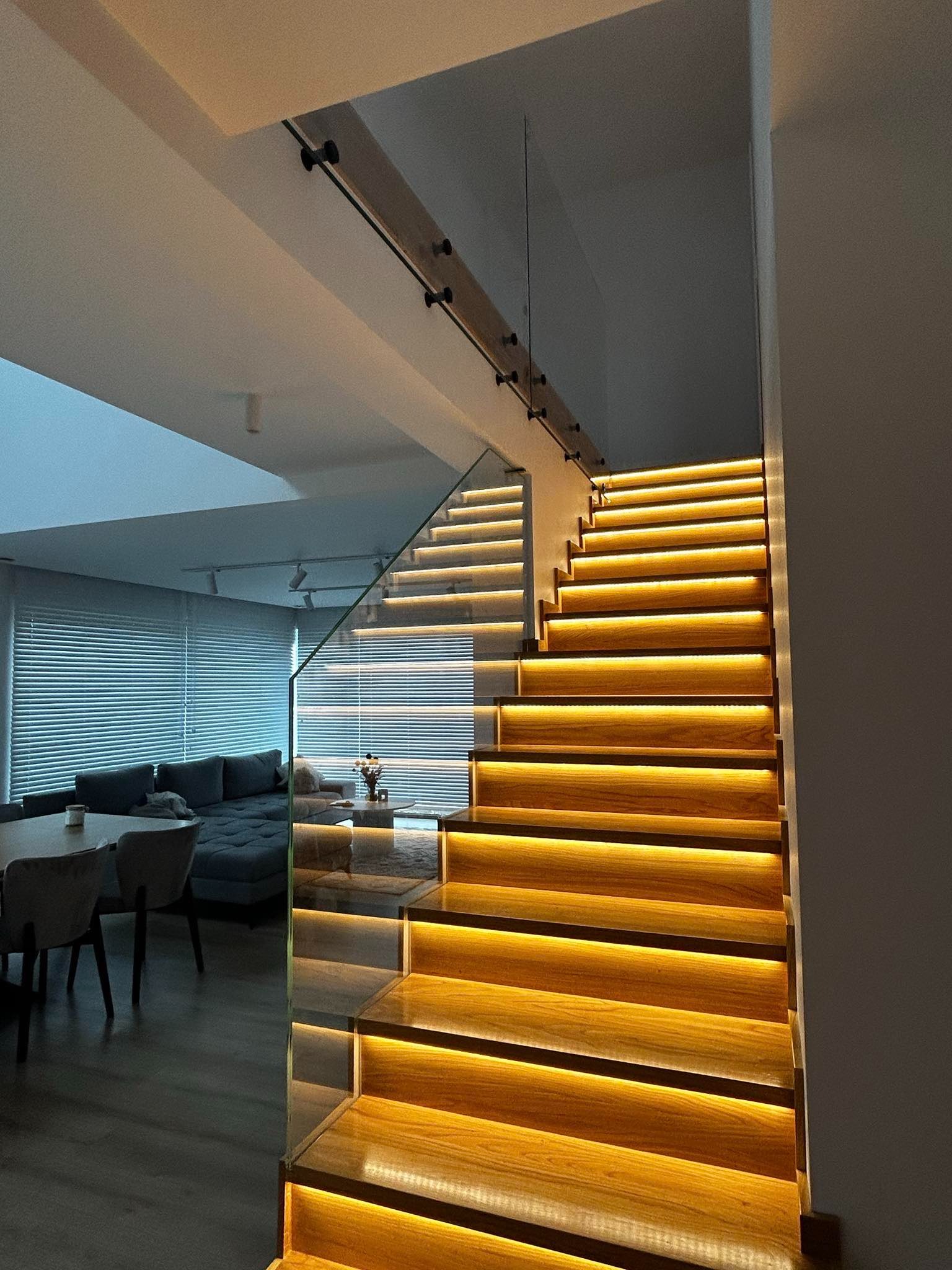 Nowoczesne schody z podświetlanymi stopniami i szklaną balustradą, widok z dołu na tle salonu z szarą sofą i żaluzjami.