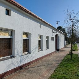 GNHome - Boczny widok odnowionego budynku z białą elewacją i brązowymi detalami, takimi jak dach, rynny i podmurówka, z widocznymi oknami z żaluzjami, położonego przy chodniku z kostki brukowej i ulicy...