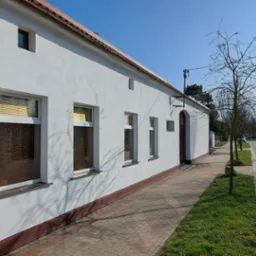 Boczny widok odnowionego budynku z białą elewacją i brązowymi detalami, takimi jak dach, rynny i podmurówka, z widocznymi oknami z żaluzjami, położonego przy chodniku z kostki brukowej i ulicy...