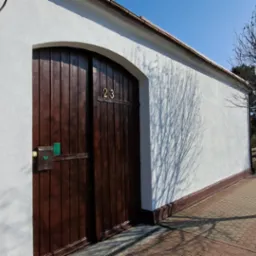 Biała elewacja budynku z ciemnobrązową, drewnianą bramą z numerem 23, widok z poziomu chodnika w słoneczny dzień, cień drzewa na ścianie.