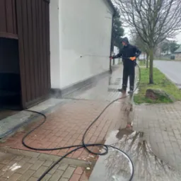 Mężczyzna w czarnym kombinezonie i pomarańczowych rękawicach czyści elewację budynku za pomocą myjki ciśnieniowej; widoczna mokra kostka brukowa i numer 23 na budynku.