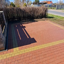 GNHome - Czerwona kostka brukowa ułożona na podjeździe z żółtymi akcentami, widoczne cienie drzew, w tle krzewy i domy pod błękitnym niebem.