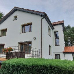 GNHome Grzegorz Nowicki - Dwupiętrowy dom z białą elewacją, brązowymi oknami i balkonem z kwiatami, otoczony zielenią. Widok z dołu, podkreślający wysokość budynku i jego otoczenie.