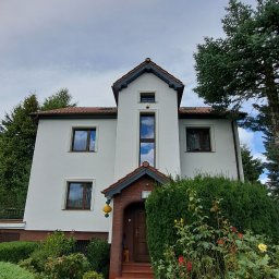 GNHome - Biały dom jednorodzinny z brązowymi oknami i ceglanym wejściem, otoczony zielenią. Widok z dołu, podkreślający wysokość budynku i jego symetryczną fasadę.