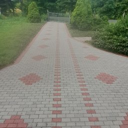 GNHome - Ułożona kostka brukowa w Trzebórzu tworząca wzór na podjeździe, prowadząca do bramy w tle. Otoczenie zielone, drzewa i krzewy.