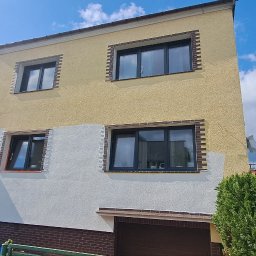 GNHome Grzegorz Nowicki - Dwukondygnacyjny dom z częściowo pomalowaną elewacją na żółto i biało, widoczne okna z ciemnymi ramami, drabina oparta o ścianę, zieleń w tle i błękitne niebo.