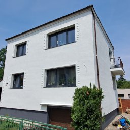 GNHome Grzegorz Nowicki - Biała, dwukondygnacyjna elewacja domu z balkonem, szarą podmurówką i ciemnymi oknami. Widok z dołu, błękitne niebo w tle. Nowoczesny styl.