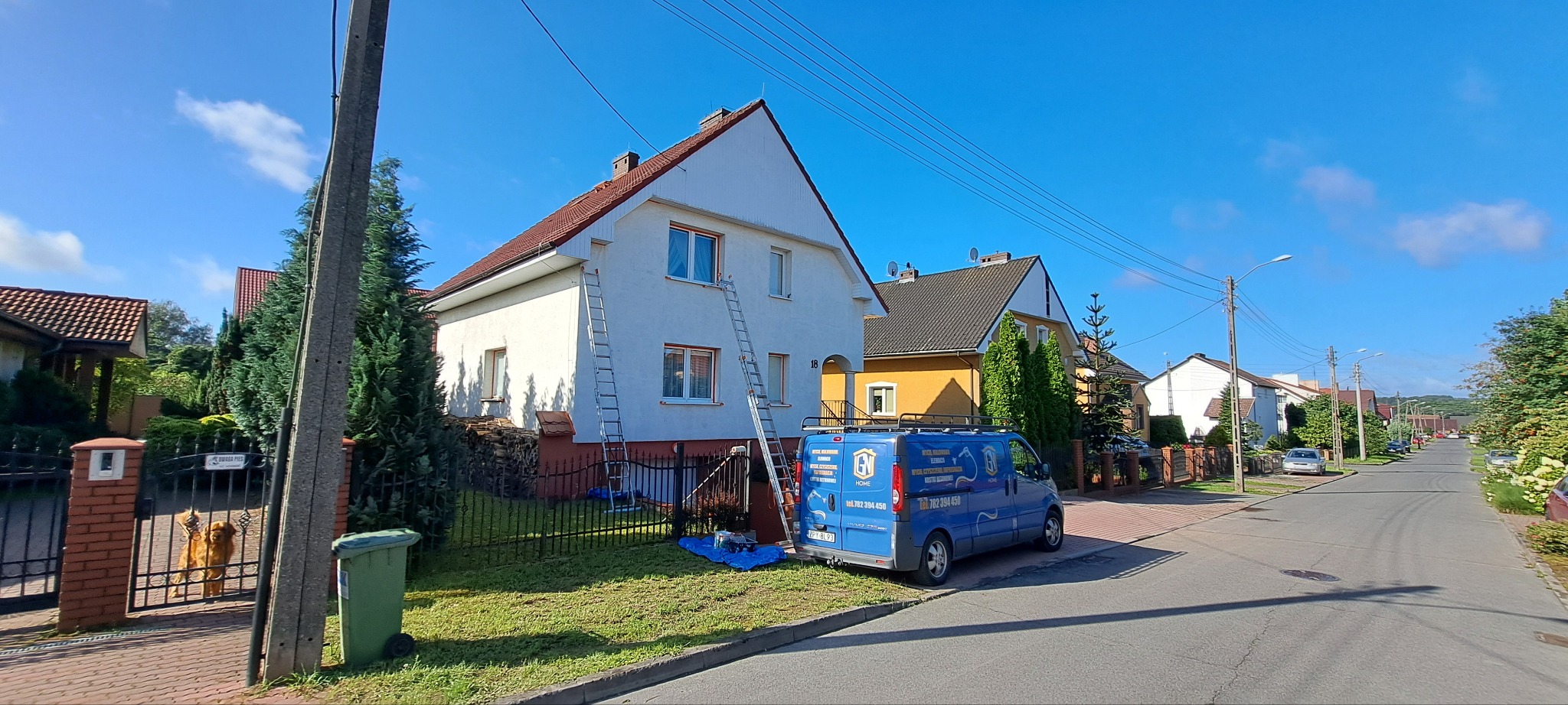 Dom jednorodzinny z drabinami oparty o elewację, sugerujący prace malarskie. Na pierwszym planie błękitny samochód dostawczy z logo firmy. Słoneczny dzień.