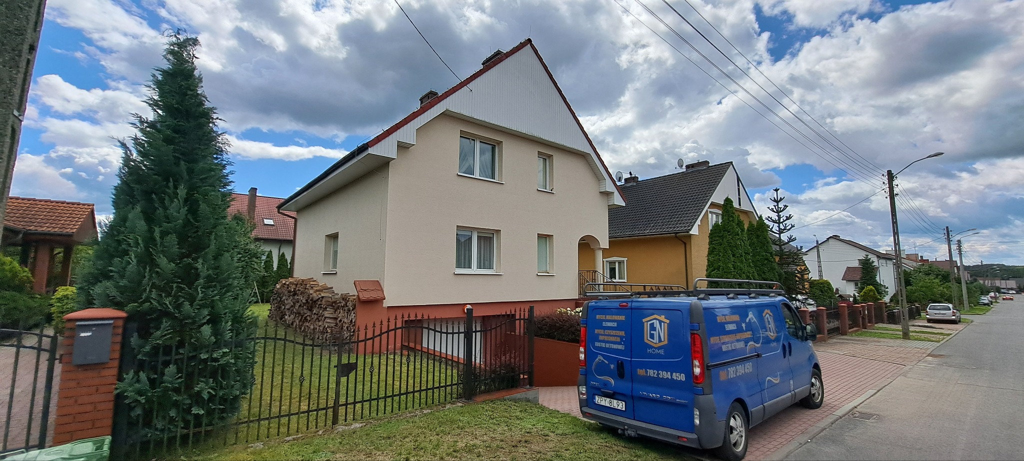 Dwupiętrowy dom z beżową elewacją, widoczny zza czarnego ogrodzenia. Na pierwszym planie zaparkowany niebieski bus z reklamą usług elewacyjnych.