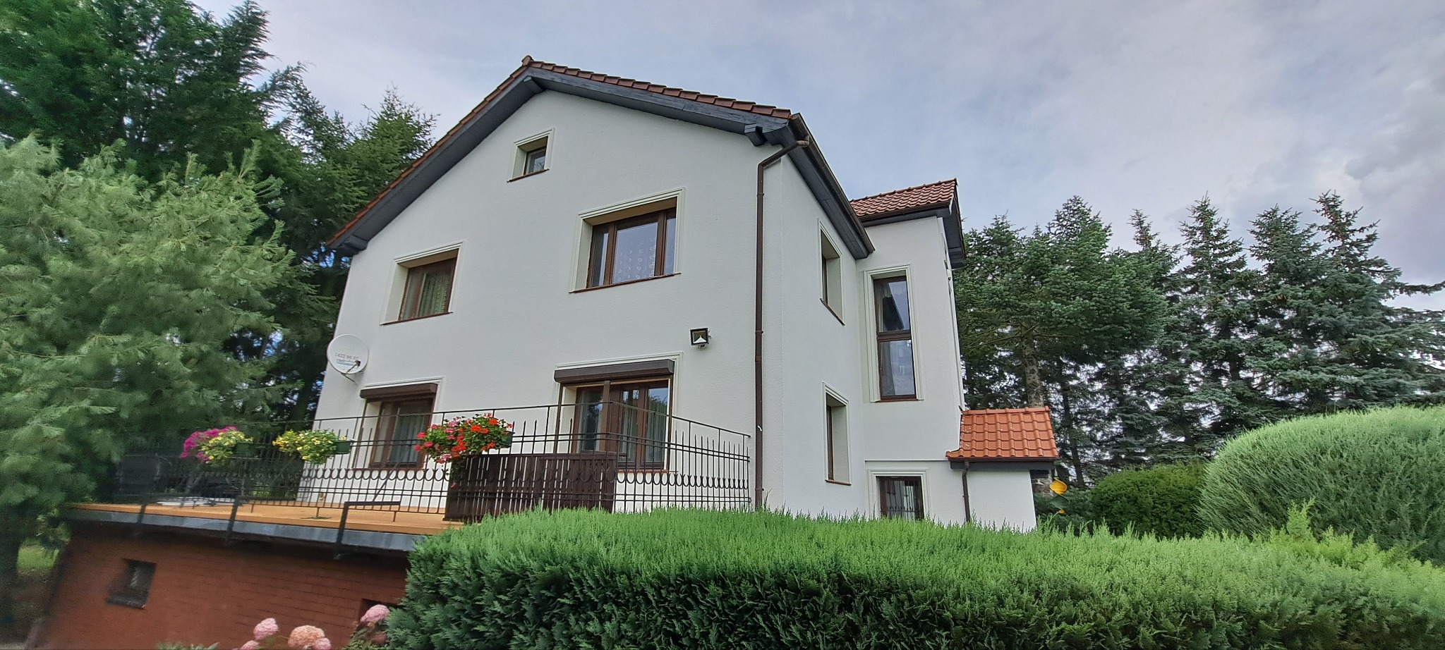 Dwupiętrowy dom z białą elewacją, brązowymi oknami i balkonem z kwiatami, otoczony zielenią. Widok z dołu, podkreślający wysokość budynku i jego otoczenie.