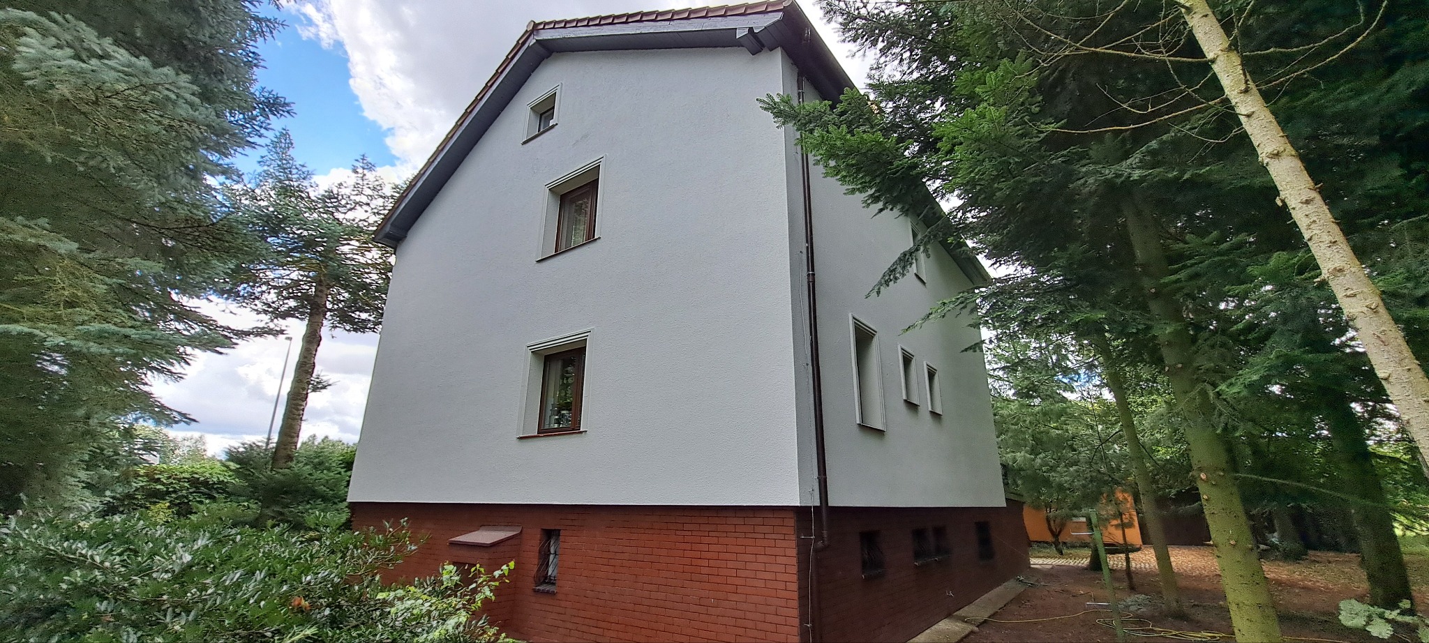 Odnowiona elewacja domu z białym tynkiem i ceglaną podmurówką, otoczona zielenią drzew. Widoczne okna z brązowymi ramami i fragment dachu. Ujęcie z dołu.