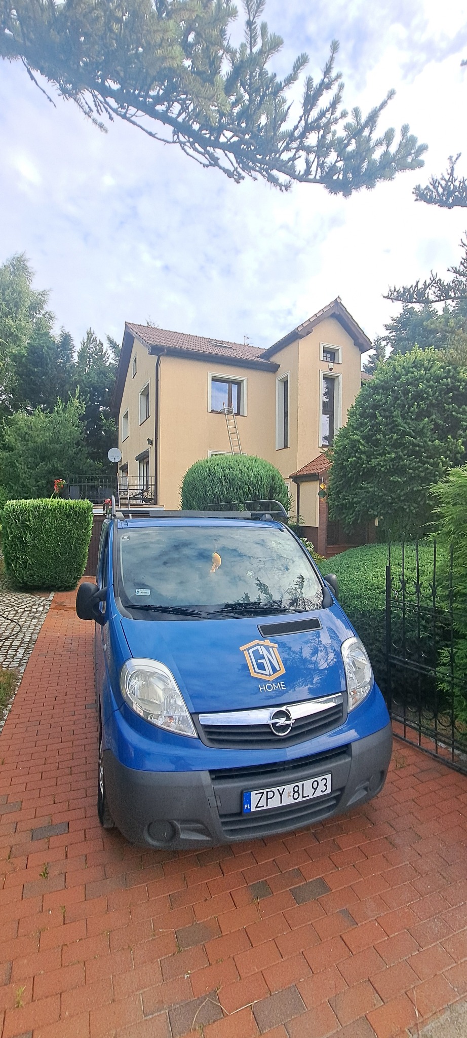 Jasna elewacja domu z drabiną opartą o ścianę, widoczna zza niebieskiego samochodu firmy z logo GN Home na tle kostki brukowej i zieleni.