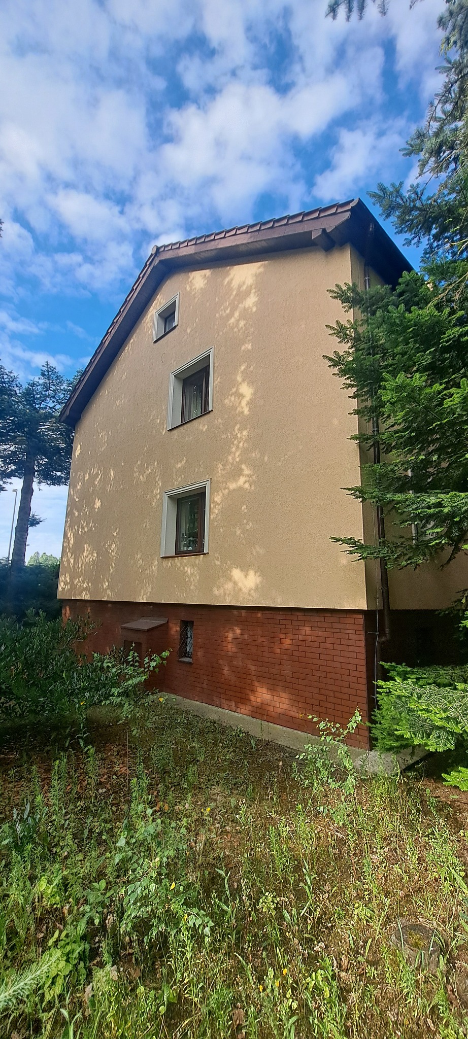 Elewacja domu jednorodzinnego w odcieniach beżu i brązu, z widocznymi oknami i fragmentem zieleni. Ujęcie z dołu, podkreślające wysokość budynku.