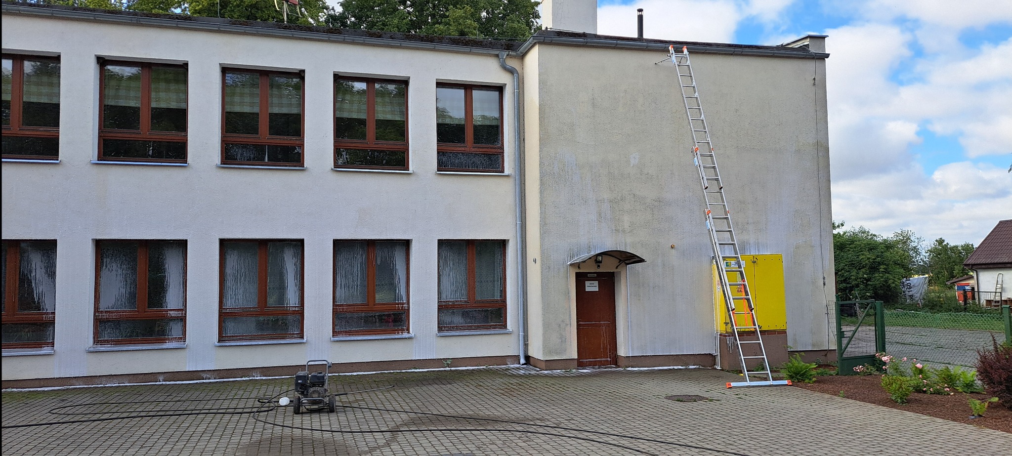 Mycie elewacji budynku z widocznym sprzętem wysokociśnieniowym i drabiną opartą o ścianę. Częściowo umyta ściana z zaciekami. Kostka brukowa na pierwszym planie.
