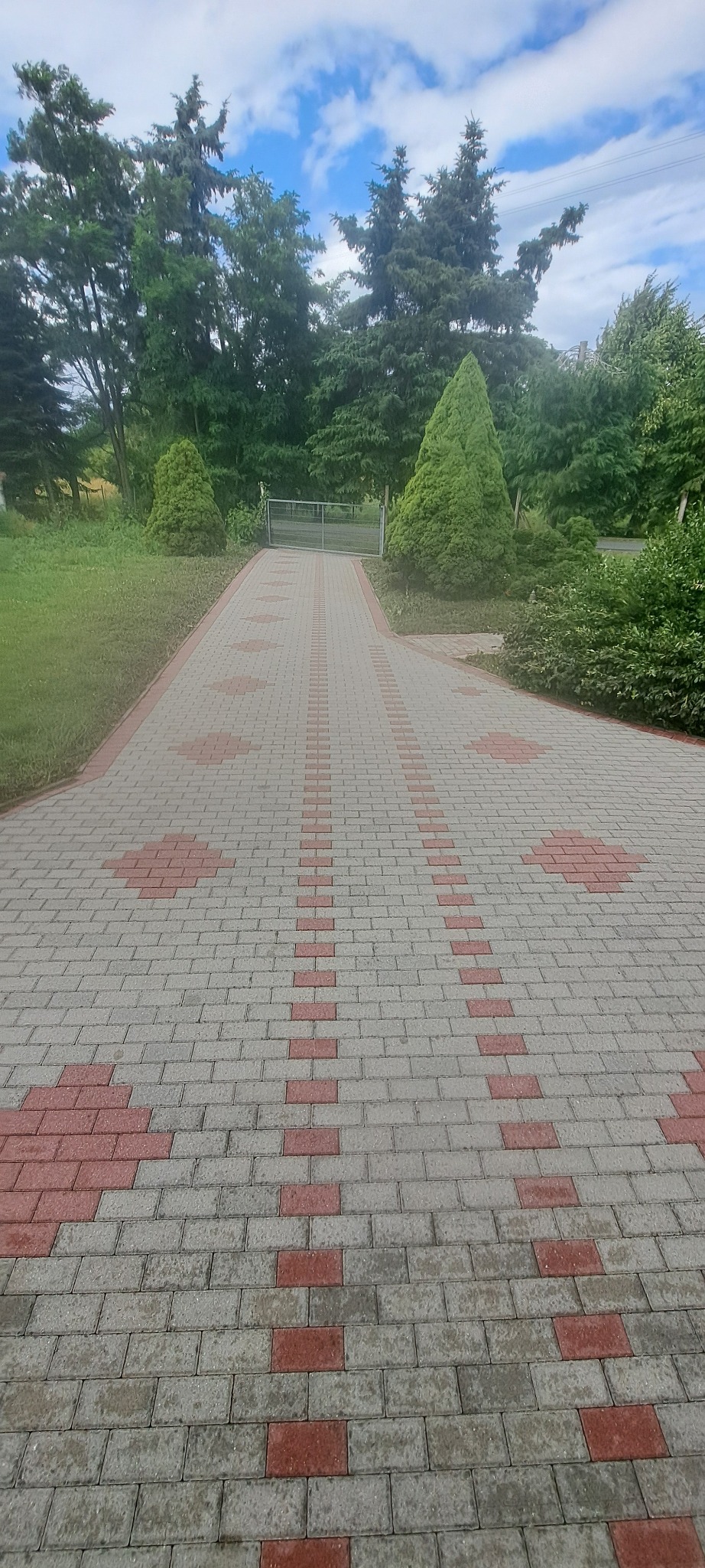 Ułożona kostka brukowa w Trzebórzu tworząca wzór na podjeździe, prowadząca do bramy w tle. Otoczenie zielone, drzewa i krzewy.