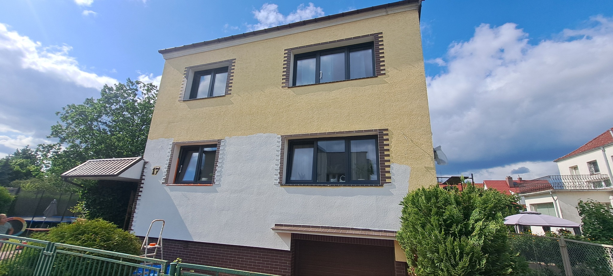 Dwukondygnacyjny dom z częściowo pomalowaną elewacją na żółto i biało, widoczne okna z ciemnymi ramami, drabina oparta o ścianę, zieleń w tle i błękitne niebo.