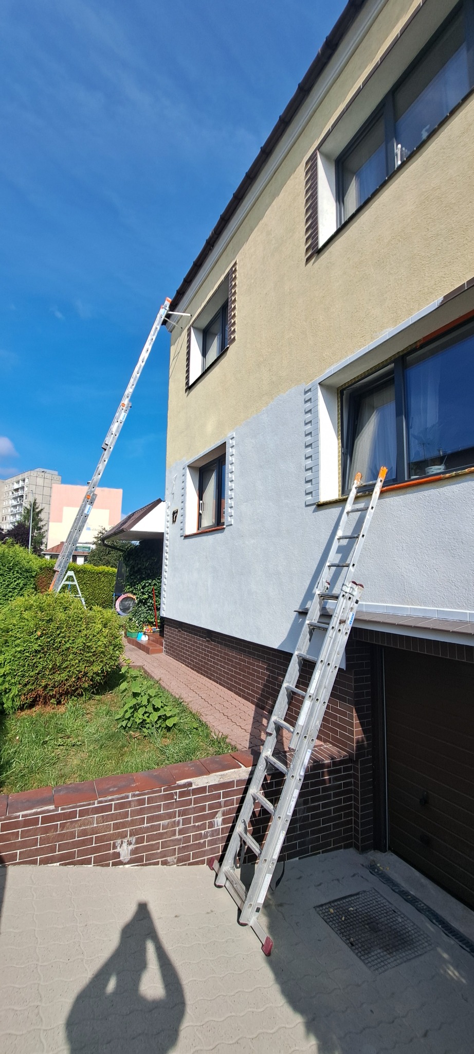 Fragment elewacji domu w trakcie odświeżania, z widocznymi drabinami i częściowo pomalowaną ścianą na szaro. Jasne, słoneczne światło.