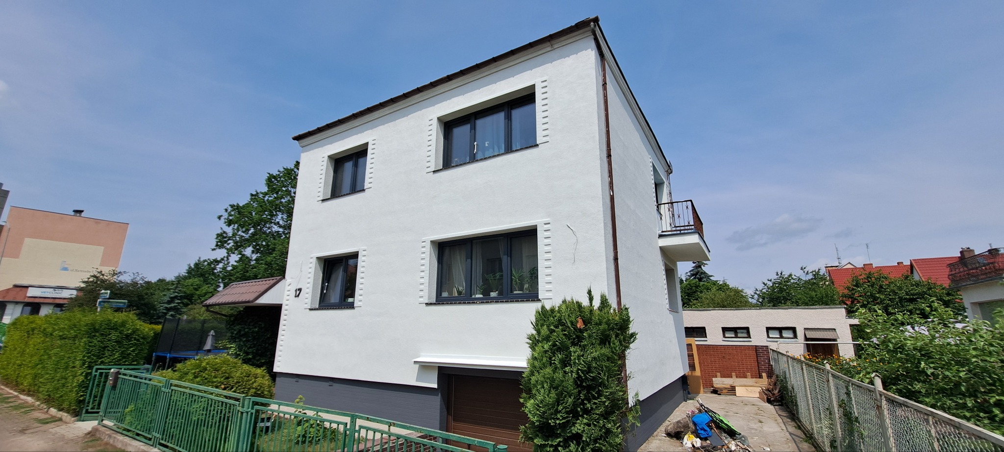 Biała, dwukondygnacyjna elewacja domu z balkonem, szarą podmurówką i ciemnymi oknami. Widok z dołu, błękitne niebo w tle. Nowoczesny styl.