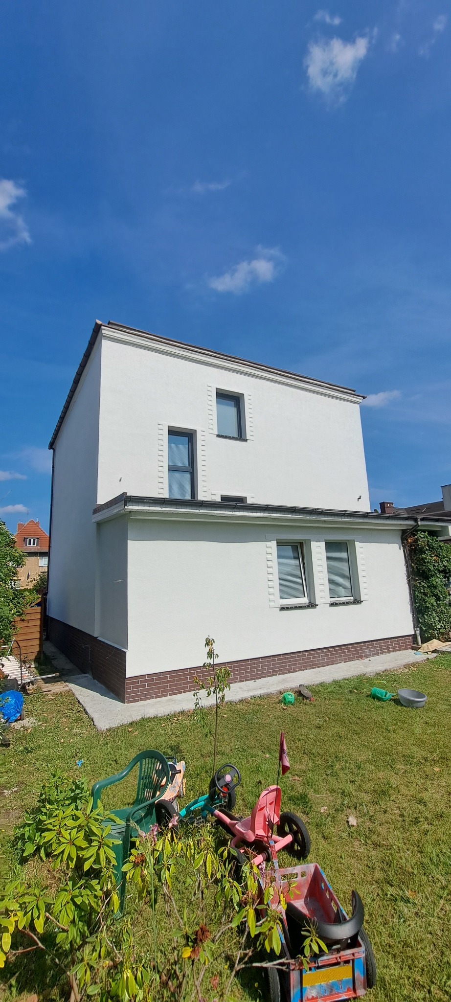 Dwukondygnacyjny dom z białą elewacją i ceglaną podmurówką, widziany z poziomu trawnika z zabawkami. Błękitne niebo w tle.