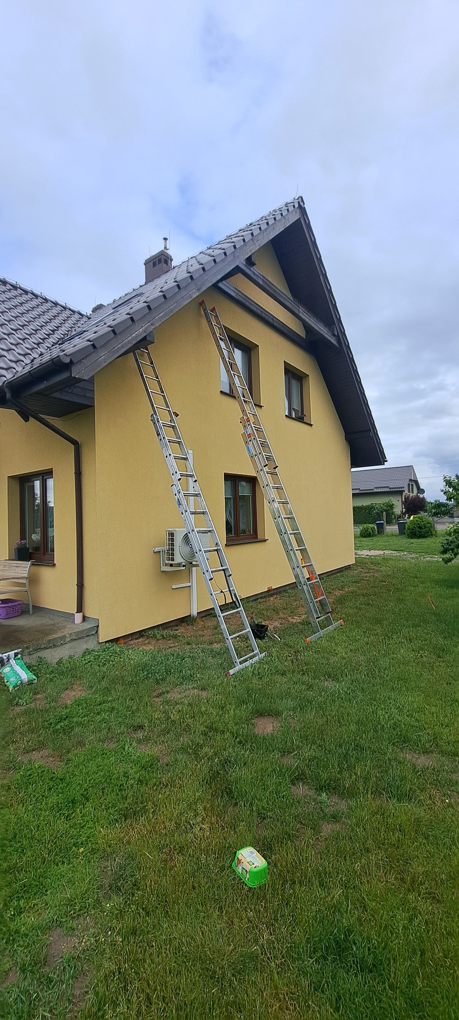Żółta elewacja domu z brązowymi oknami i dwiema rozstawionymi drabinami. Widoczne ślady po malowaniu. Na trawie leży zielone pudełko.