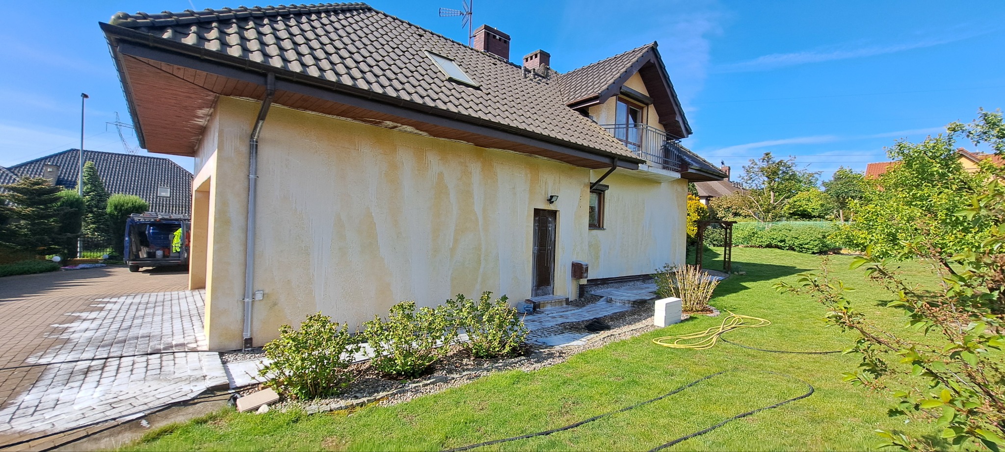 Boczny widok domu z elewacją w trakcie mycia. Widoczne ślady po czyszczeniu na ścianie i kostce brukowej. Na podjeździe zaparkowany bus z narzędziami.