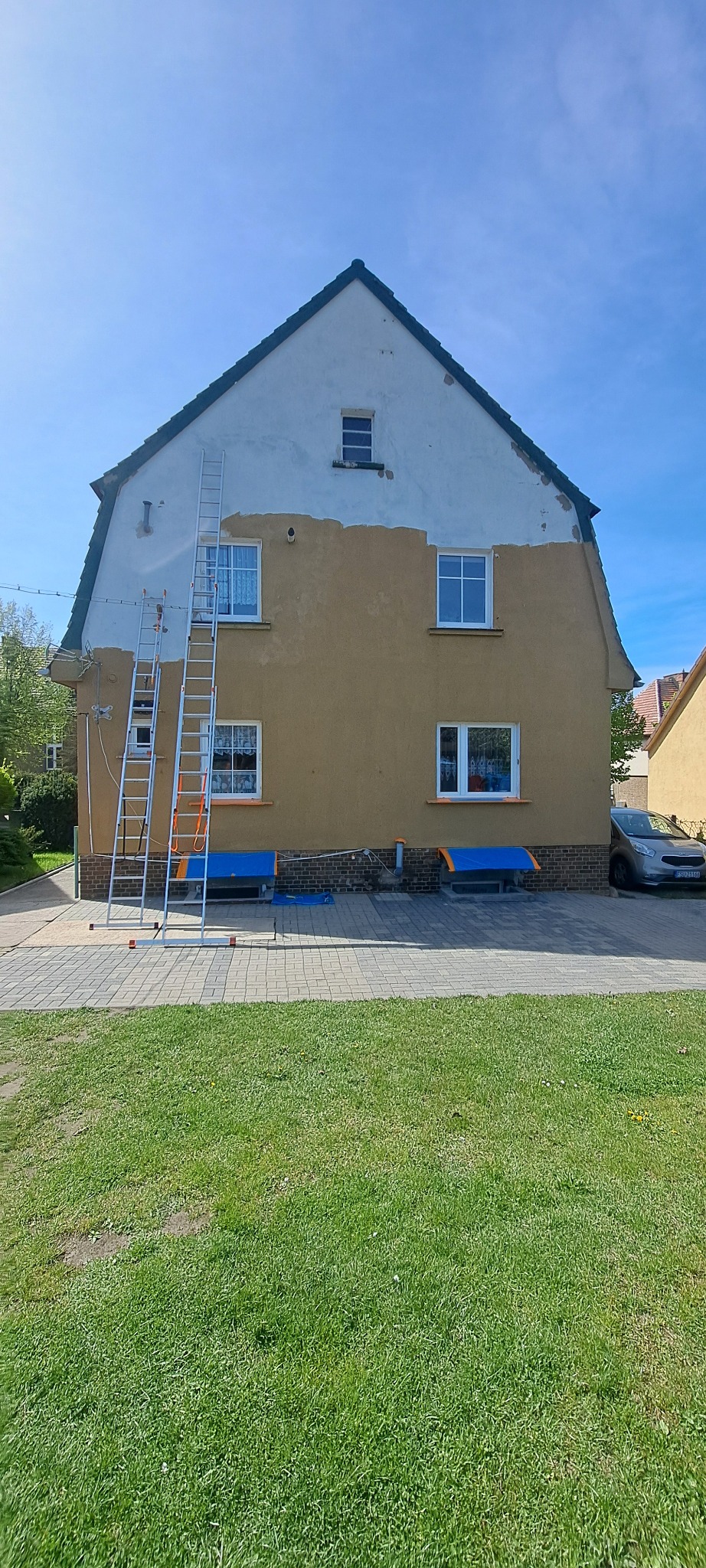 Dwukondygnacyjny dom z częściowo pomalowaną elewacją na brązowo, z widocznymi drabinami i zabezpieczeniami okien. Prace w toku, widok z poziomu trawnika.