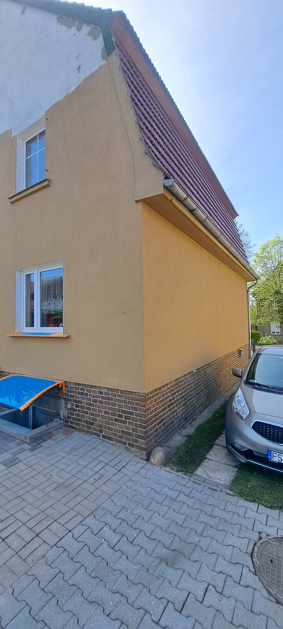 Fragment domu z ocieploną, beżową elewacją i czerwoną dachówką, widoczny z boku. Część elewacji wymaga poprawek. Obok zaparkowany samochód.