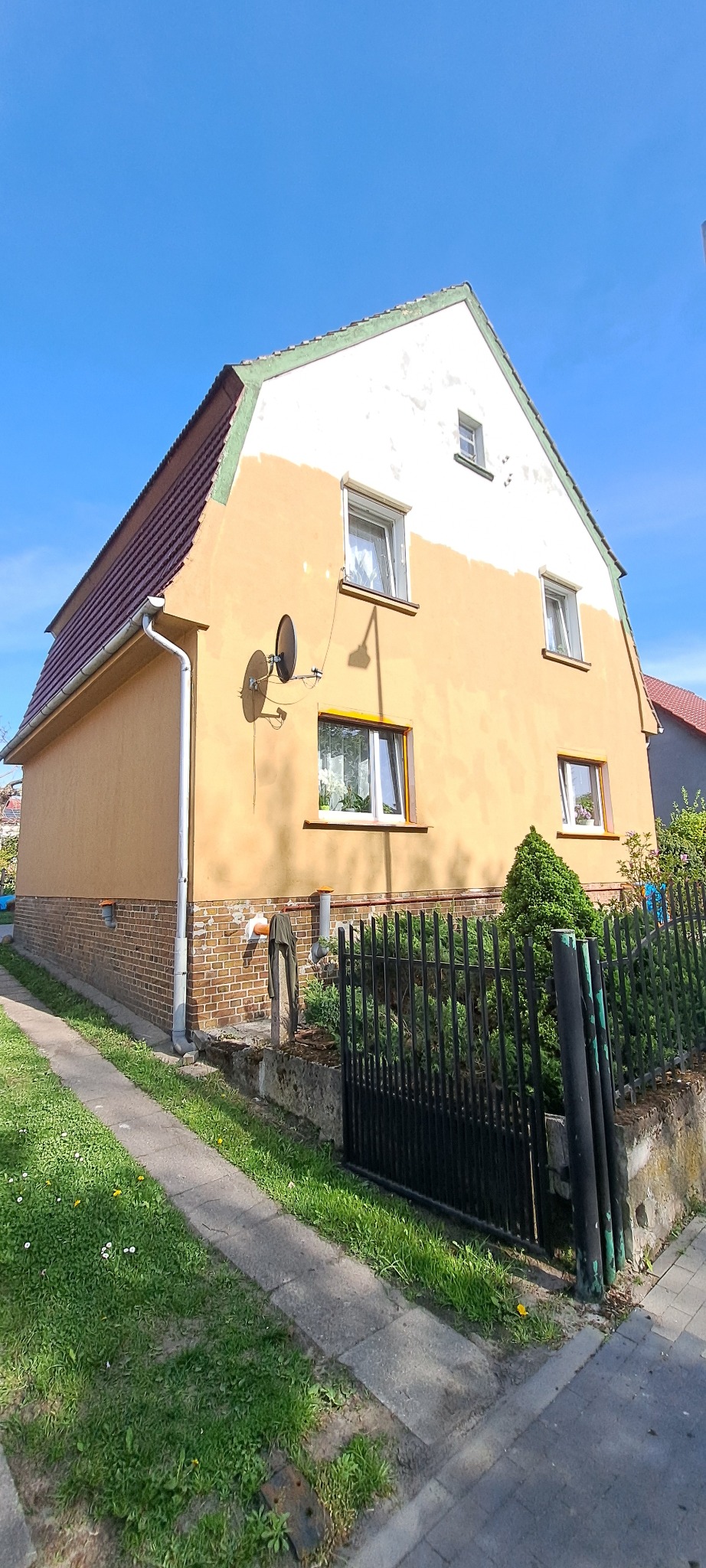 Dom jednorodzinny z częściowo pomalowaną elewacją na żółto i biało, widoczne anteny satelitarne, zielony trawnik i czarne ogrodzenie. Słoneczny dzień.