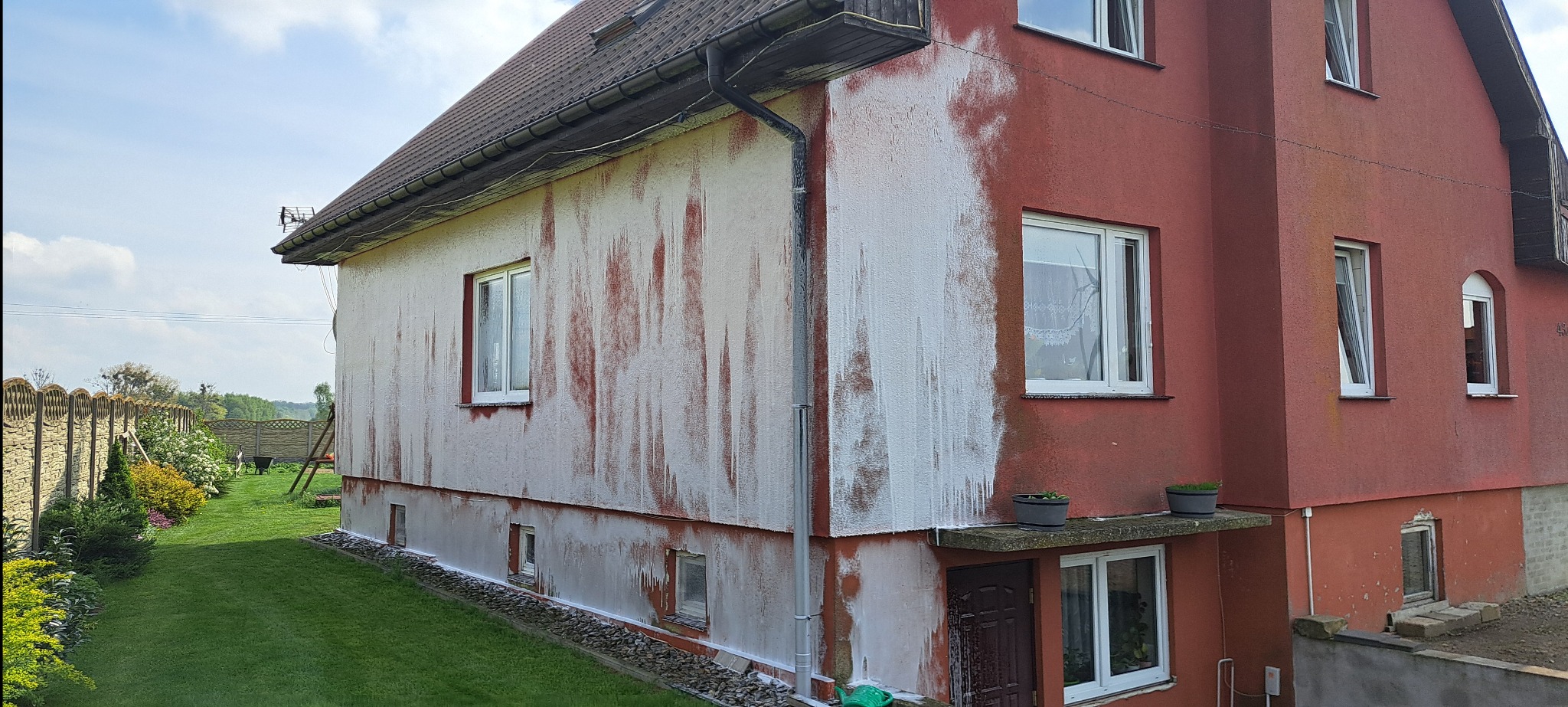 Dom z częściowo umytą elewacją, ukazujący kontrast przed i po czyszczeniu. Widoczne zacieki i różnica w kolorze. Ujęcie z ogrodu, zieleń trawy i roślin w tle.