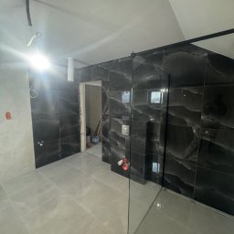 Ciemna, marmurkowa glazura ścienna w łazience z kabiną prysznicową typu walk-in. Widoczne szare płytki podłogowe i fragment instalacji elektrycznej.