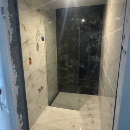 Wykończona kabina prysznicowa z marmurowymi płytkami w odcieniach bieli i czerni. Widoczne elementy armatury i oświetlenie sufitowe. Częściowo widoczne surowe wykończenie ściany.