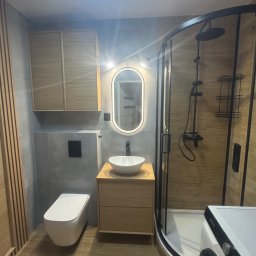 Nowoczesna łazienka z kabiną prysznicową typu walk-in, podświetlanym lustrem i drewnianymi akcentami. Uwagę zwraca minimalistyczna toaleta i umywalka nablatowa.