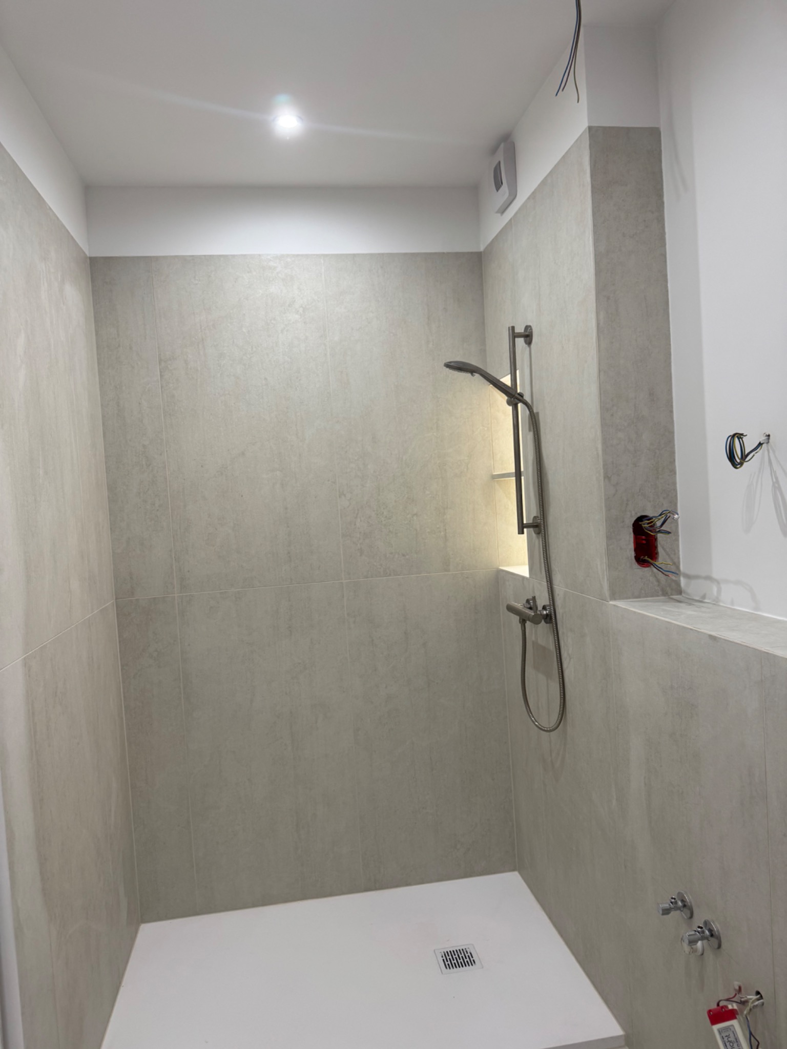 Wykończona kabina prysznicowa z beżowymi płytkami, białą brodzikiem i chromowaną armaturą. Widoczne niedokończone prace elektryczne. Minimalistyczny design.