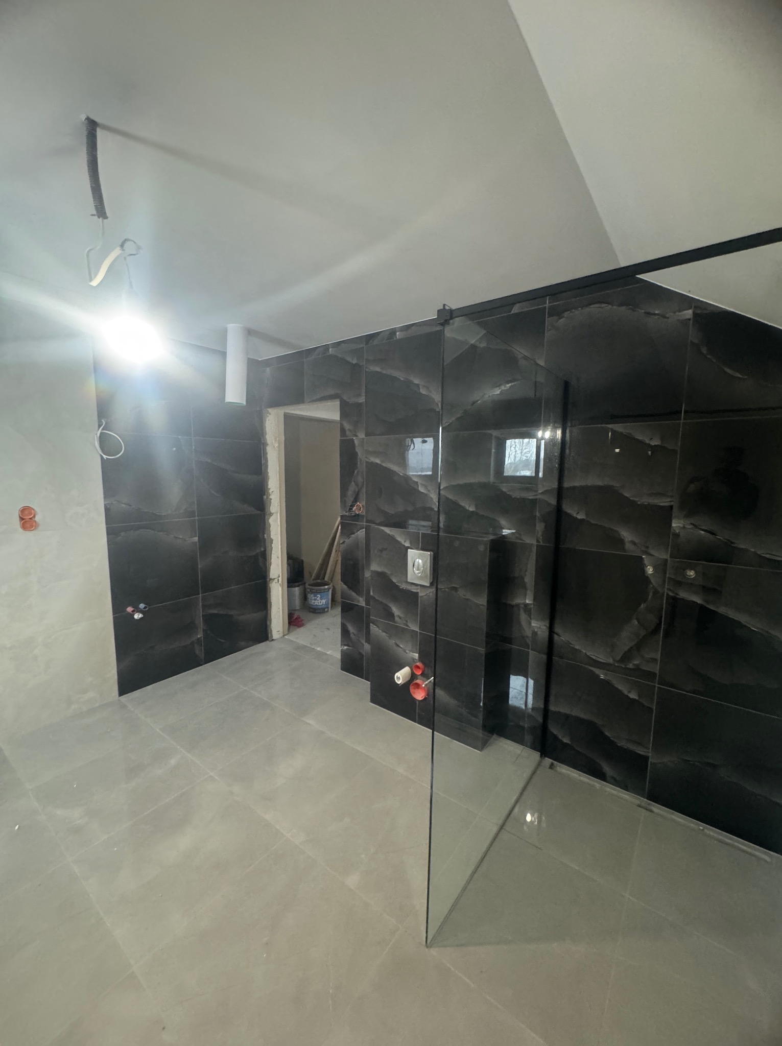 Ciemna, marmurkowa glazura ścienna w łazience z kabiną prysznicową typu walk-in. Widoczne szare płytki podłogowe i fragment instalacji elektrycznej.