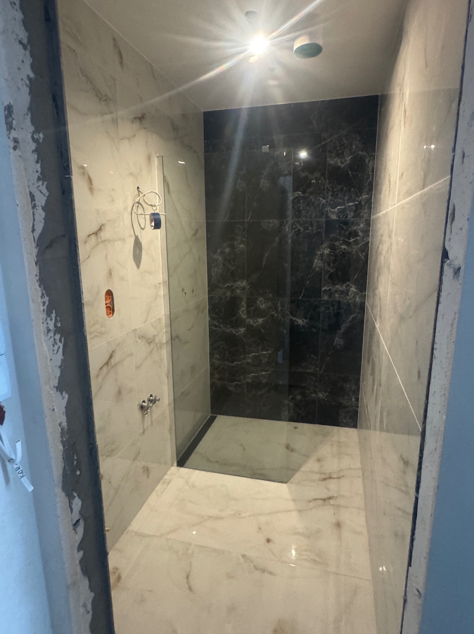 Wykończona kabina prysznicowa z marmurowymi płytkami w odcieniach bieli i czerni. Widoczne elementy armatury i oświetlenie sufitowe. Częściowo widoczne surowe wykończenie ściany.