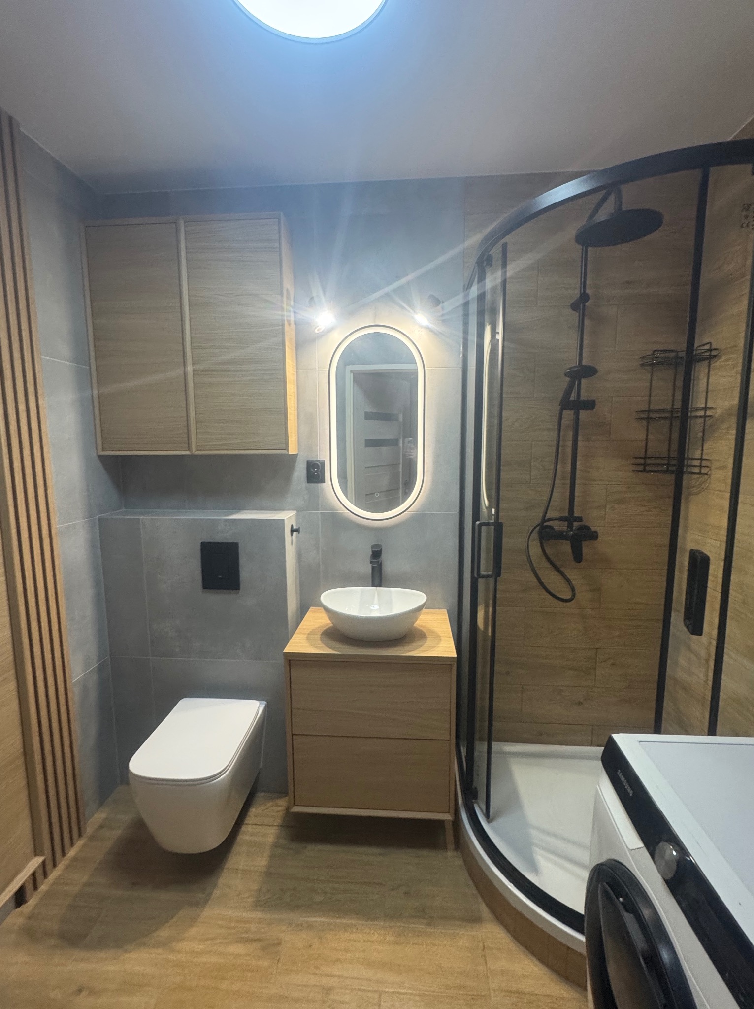 Nowoczesna łazienka z kabiną prysznicową typu walk-in, podświetlanym lustrem i drewnianymi akcentami. Uwagę zwraca minimalistyczna toaleta i umywalka nablatowa.