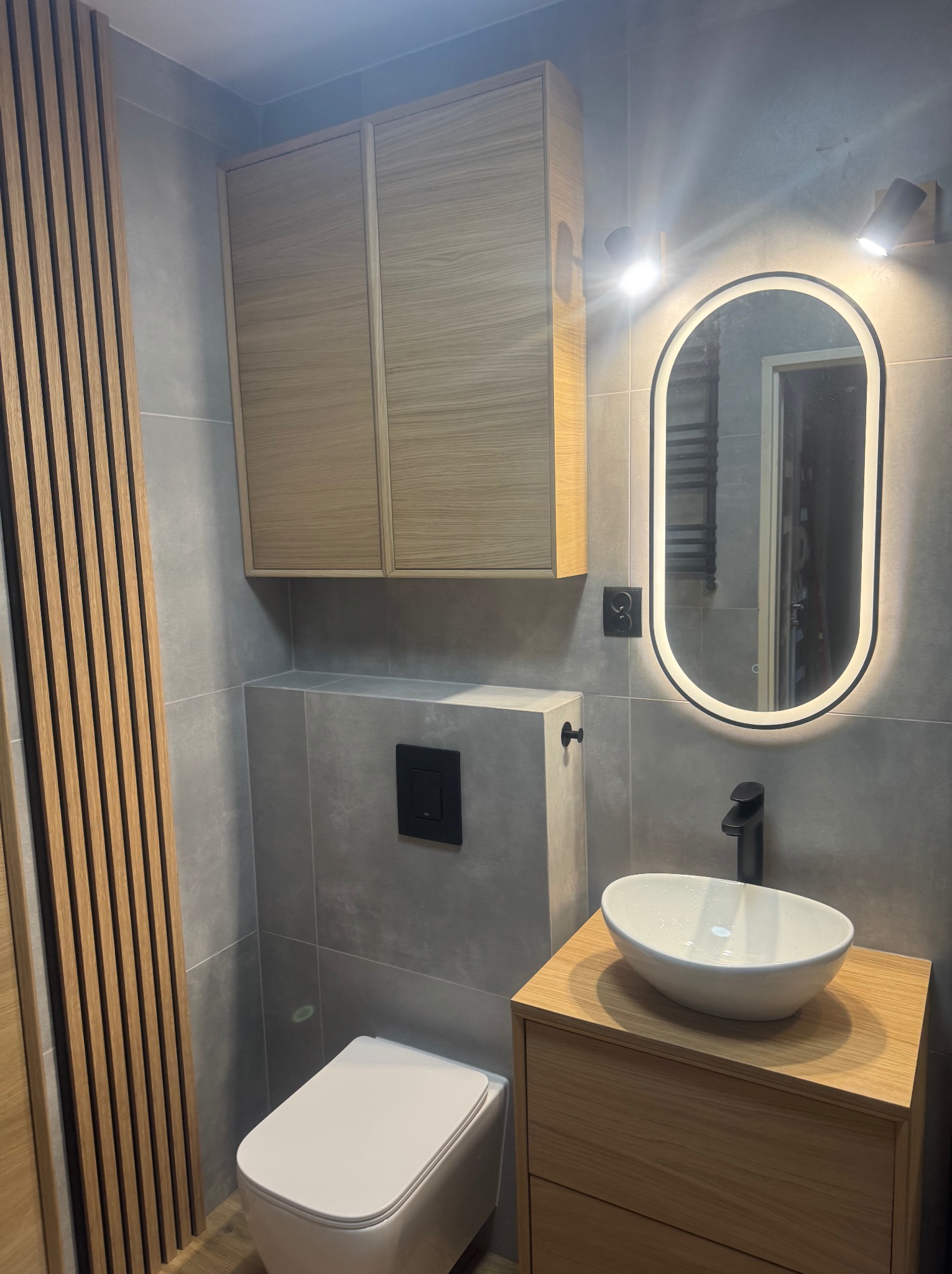 Nowoczesna łazienka w szarościach z drewnianymi akcentami: szafka pod umywalką, wisząca szafka i dekoracyjne lamele. Owalne lustro z podświetleniem LED.