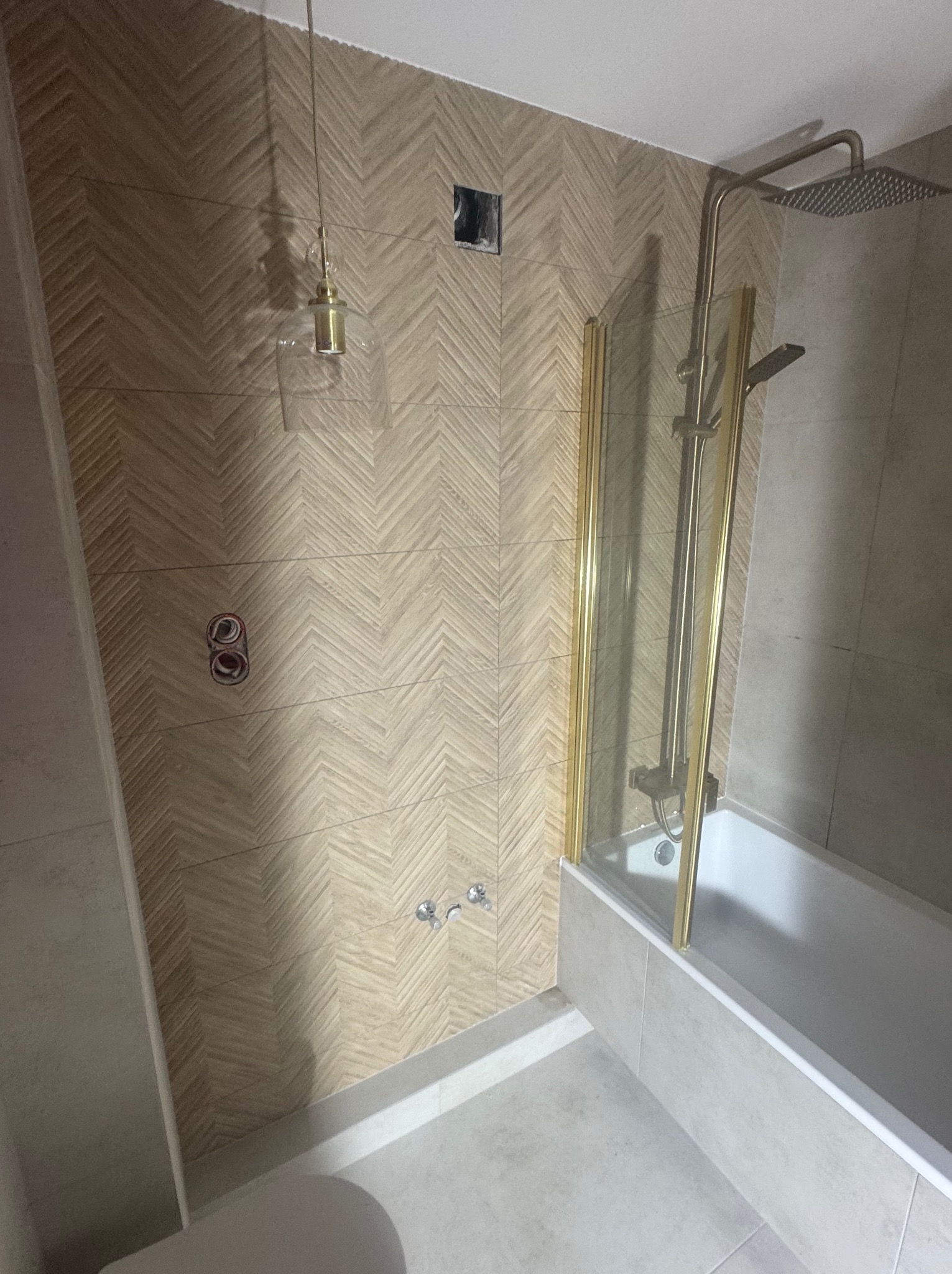 Wykończona łazienka z beżowymi płytkami ułożonymi w jodełkę, złotą armaturą i szklanym parawanem nawannowym. Widoczne przyłącza i lampa wisząca.